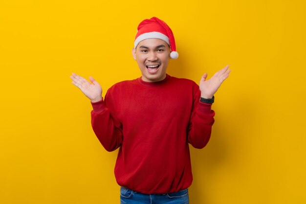 Surprised young Asian man in Santa hat raising hands and looking at camera with excitement expression on yellow studio background celebration Christmas holiday and New Year concept