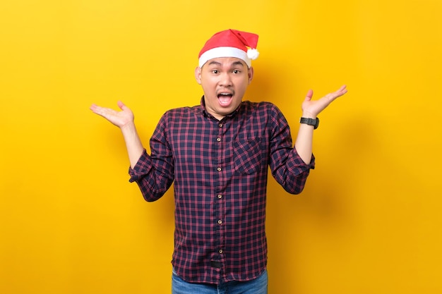 Surprised young Asian man in Santa hat raising hands and looking at camera with excitement expression on yellow studio background Happy New Year 2023 celebration merry holiday concept