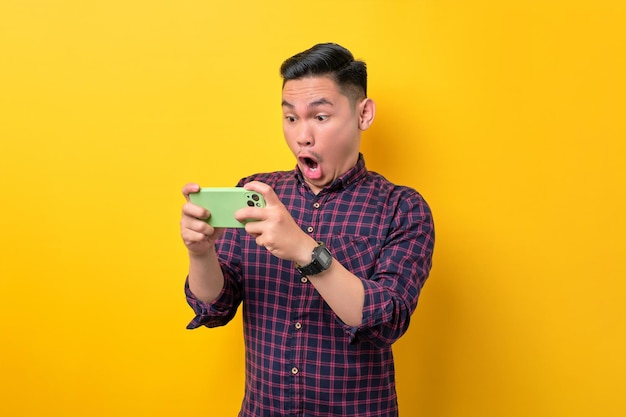 Surprised young Asian man using smartphone for playing mobile game isolated over yellow background