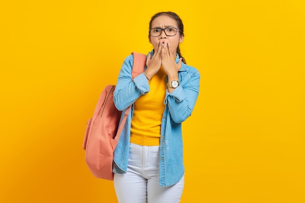 Surprised young Asian student in denim clothes with backpack covering mouth with hands isolated on yellow background Education in high school university college concept