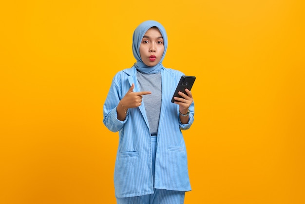 Surprised young Asian woman pointing at smartphone in hands isolated over yellow background