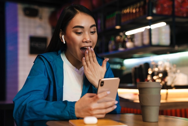 surprised young asian woman using mobile phone and wireless earphones while sitting in cafe