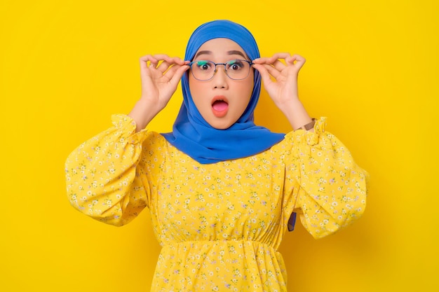 Surprised young beautiful Asian Muslim woman touching glasses with open mouth isolated over yellow background