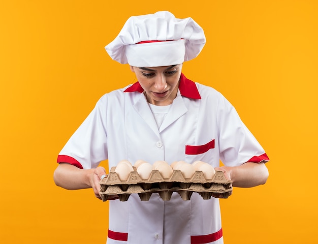 Surprised young beautiful girl in chef uniform holding and looking at batch of eggs isolated on orange wall