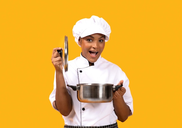 Surprised young black chef woman opening saucepan and looking at camera