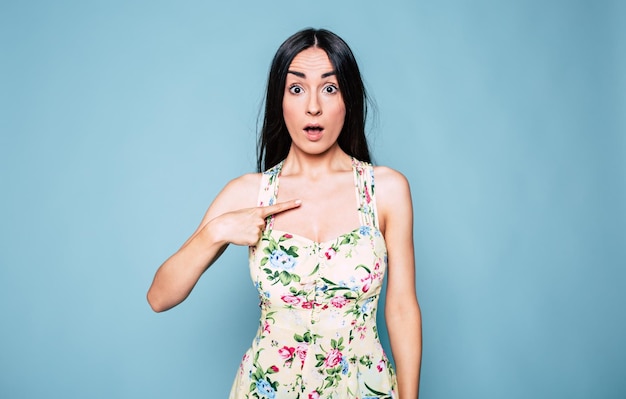 Surprised young brunette woman in the dress looks shocked on camera isolated on blue background