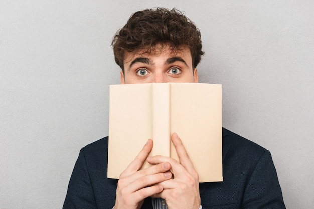 Surprised young businessman wearing suit standing isolated over gray, covering his face with a book