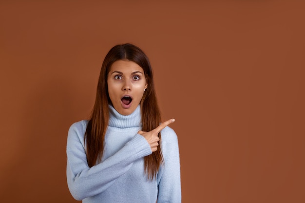 Surprised young european woman wearing light blue sweater points index finger sideways on copy space