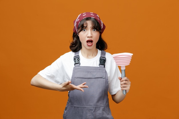 Surprised young female cleaner wearing uniform and bandana showing plunger pointing at it with hand looking at camera isolated on orange background