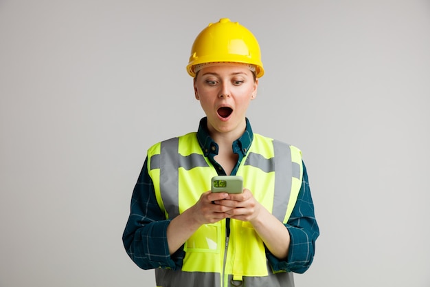 Surprised young female construction worker wearing safety helmet and safety vest holding and looking at mobile phone 