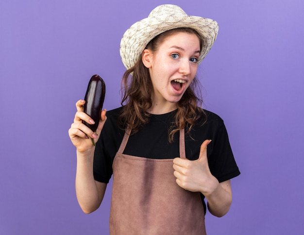 Surprised young female gardener wearing gardening hat holding eggplant showing thumb up isolated on blue wall