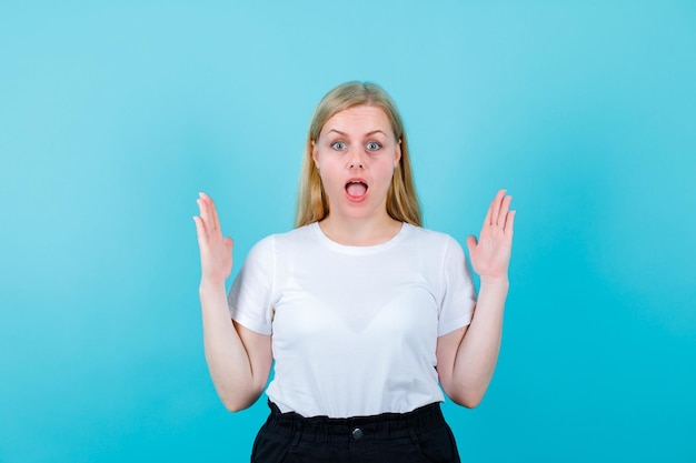 Surprised young girl is looking at camera by raising up her hands on blue background