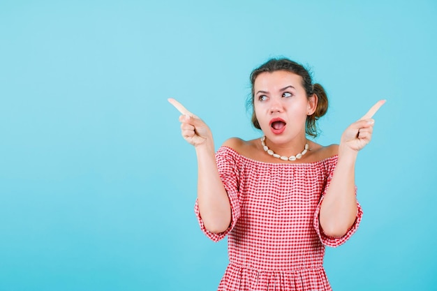 Surprised young girl is looking left and pointing up with forefingers on blue background