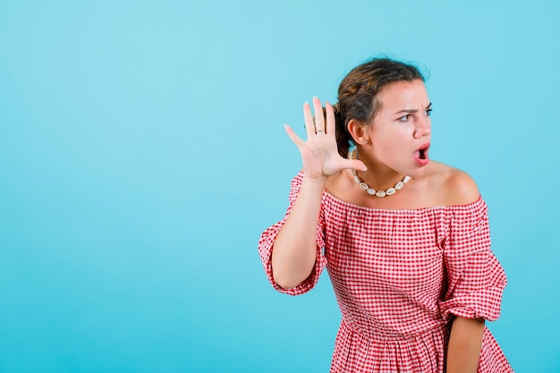 Surprised young girl is trying to hear by holding hand near ear on blue background