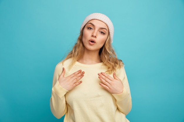 Surprised young girl wearing winter hat pointing at herself with hands 