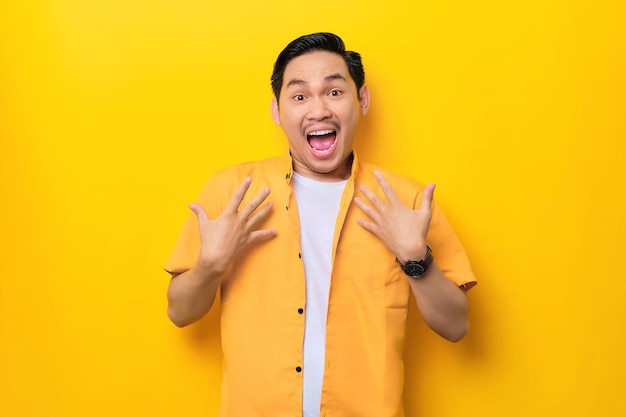 Surprised young handsome Asian man keeps hands on chest opened mouth isolated on yellow background