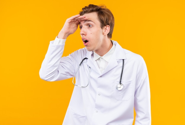 Surprised young male doctor wearing medical robe with stethoscope looking at distance with hand 