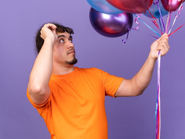 Surprised young man wearing glasses holding and looking at balloons 