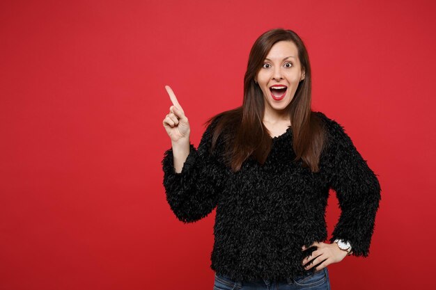 Surprised young woman in black fur sweater keeping mouth wide open, pointing index finger aside isolated on bright red wall background. People sincere emotions, lifestyle concept. Mock up copy space.