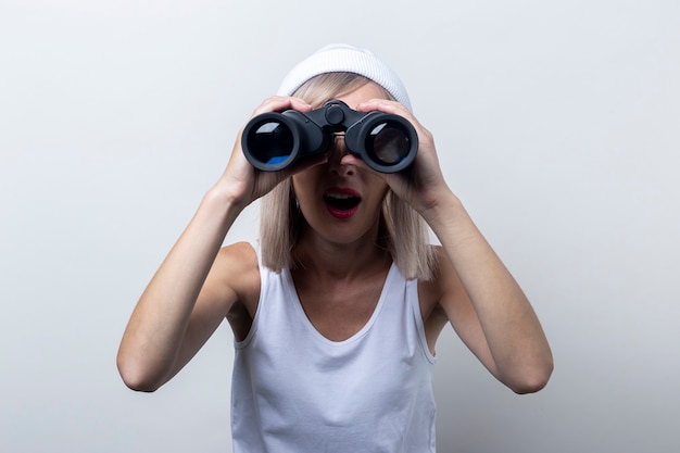 Surprised young woman looking through binoculars on a light background.