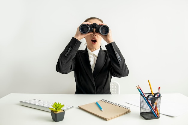 Surprised young woman looking through binoculars while sitting at desk at workplace