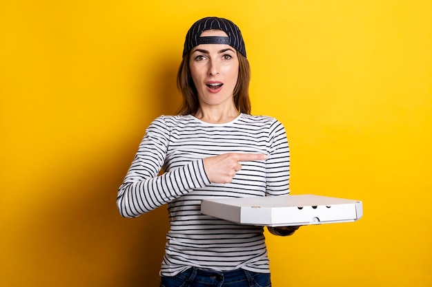 Surprised young woman pizza delivery man points finger to the side while holding package with hot pizza on yellow background