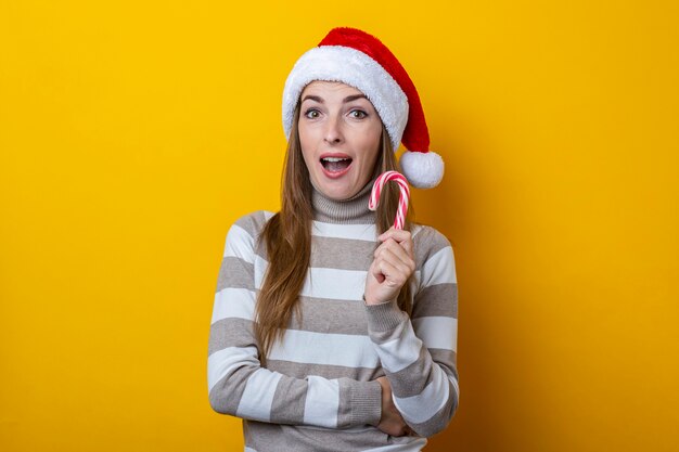 Surprised young woman in Santa Claus hat with Christmas candy on a yellow background.