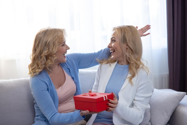 Surprised young woman seated on the sofa receiving a present from her joyful female parent