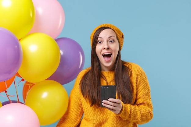 Surprised young woman in sweater hat posing isolated on blue background. Birthday holiday party, people emotions concept. Mock up copy space. Celebrating hold colorful air balloons using mobile phone.