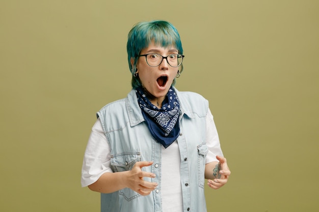 Surprised young woman wearing glasses bandana on neck keeping hands in air looking at camera isolated on olive green background