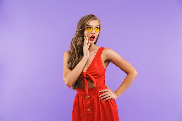 Photo surprised young woman wearing red dress and sunglasses wondering and touching cheek