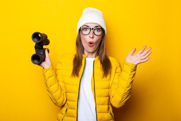 Surprised young woman in yellow jacket with binoculars on yellow surface