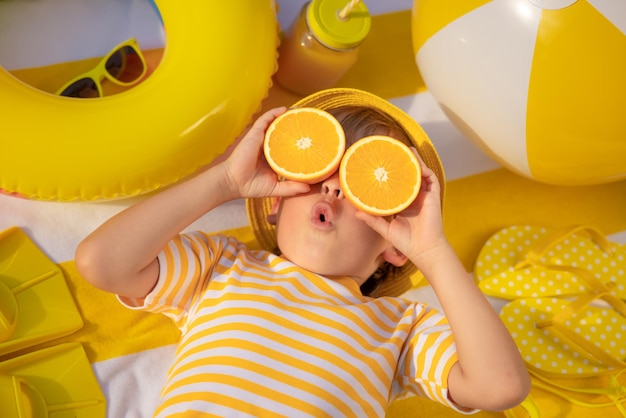 Surprized child holding slices of orange fruit like sunglasses Kid wearing striped yellow tshirt lying on beach towel Healthy eating and summer vacation concept