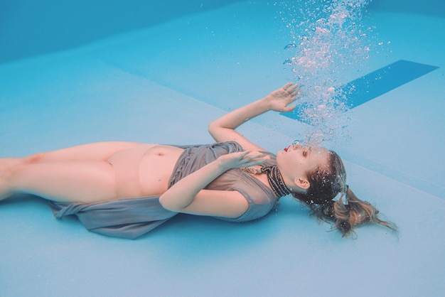 surreal art portrait of young woman in grey dress and beaded scarf underwater in the swimming pool