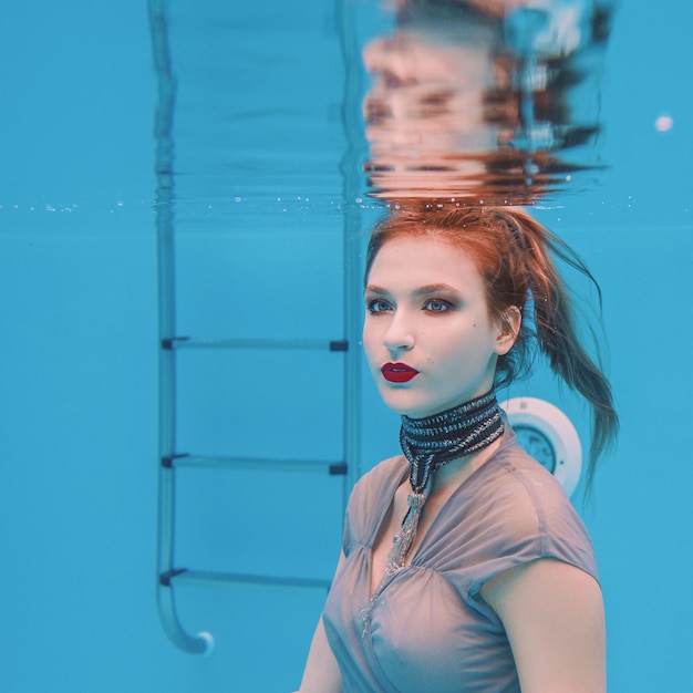 surreal art portrait of young woman in grey dress and beaded scarf underwater in the swimming pool