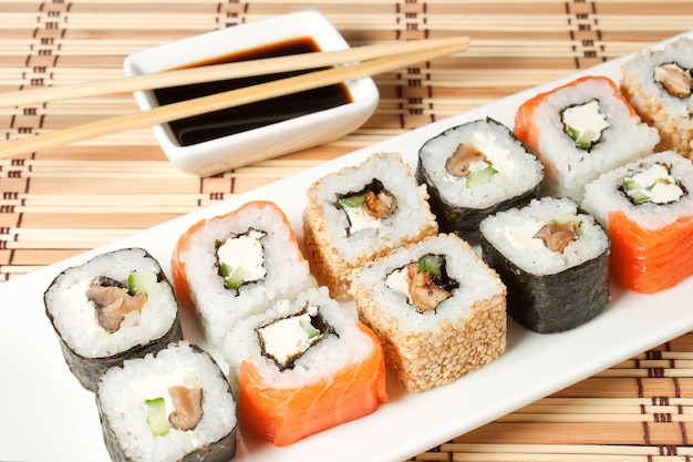 Sushi assortment on white plate, with soy sauce over bamboo background.