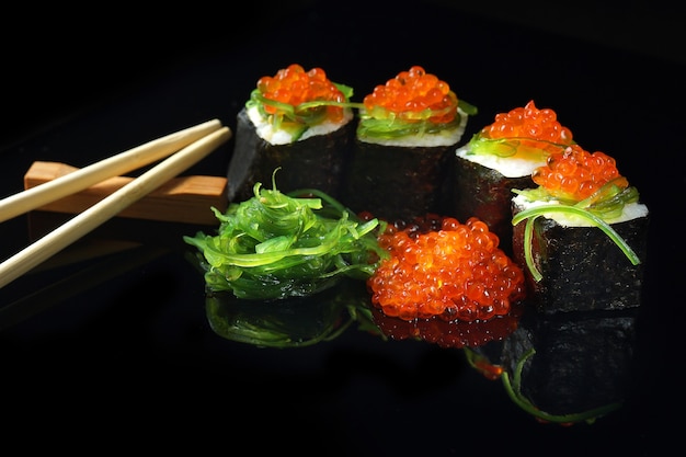 Sushi with caviar and algae salad next to chopsticks on a black with reflection.