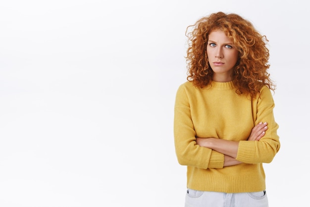 Suspicious alarmed and distressed redhead curly girlfriend in yellow sweater frowning and squinting as stare with disbelief cross arms over chest dont trust person white background