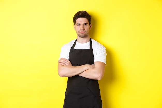 Suspicious barista squinting, cross arms on chest and looking at something with disbelief, standing over yellow wall