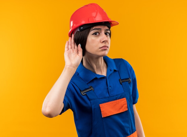 Suspicious looking at camera young builder woman in uniform showing listen gesture 