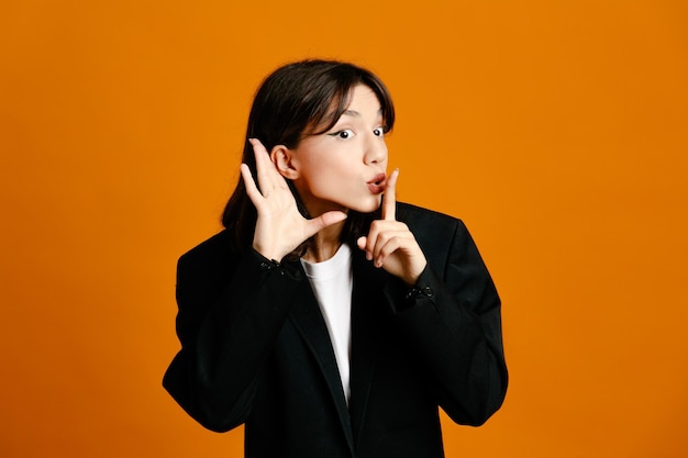 suspicious showing listen gesture young beautiful female wearing black jacket isolated on orange background