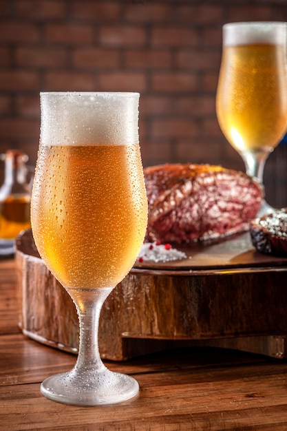 Sweaty cold tulipa glass of beer with grilled sliced cap rump steak on wooden cutting board (Brazilian picanha).