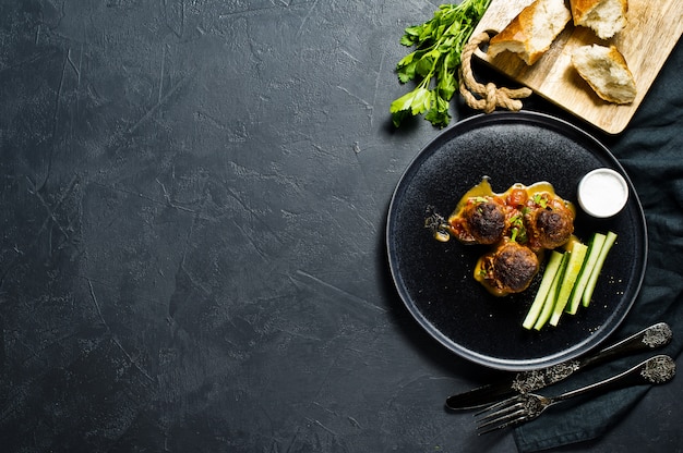 Swedish meatballs in tomato sauce on a black plate, cucumbers, salt, bread. 