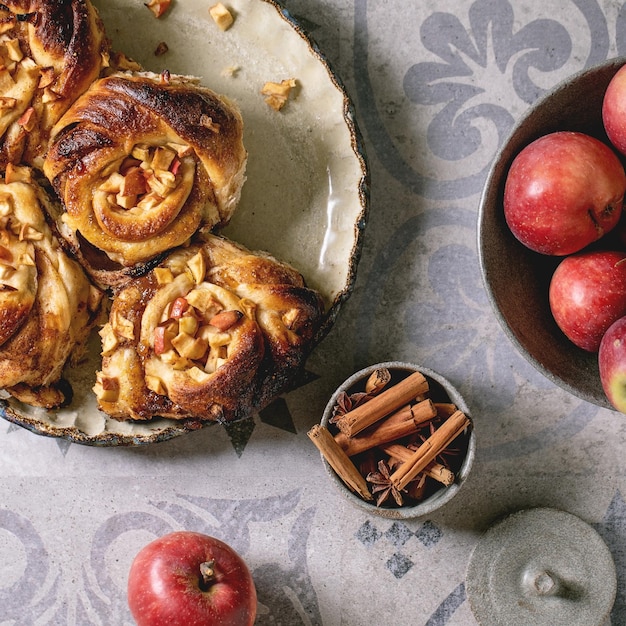 Sweet apple sticky cinnamon rolls buns start to eating in round baking tray standing with gardening apples and cinnamon sticks on ornate ceramic tile background Flat lay Square image