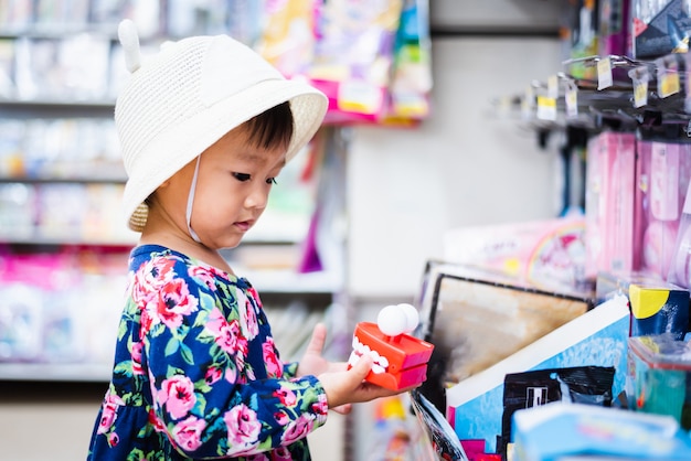 Sweet Asian girl shopping in mini mart with basket, looking at small toy in her hand