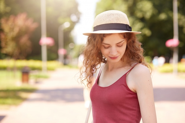 Sweet attractive young lady standing in middle of park, looking aside, enjoying time in fresh air, wearing casual summer clothes