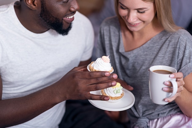 Sweet breakfast. Delighted international young couple eating cupcakes and drinking tea while enjoying the morning.