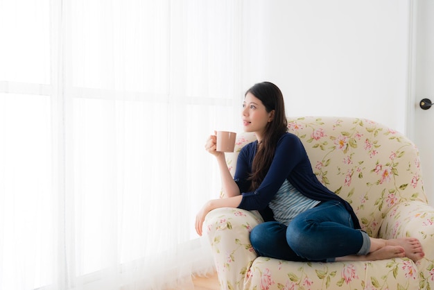 sweet calm female student holding cup drinking hot coffee and looking at window daydreaming thinking sitting on living room sofa at home.