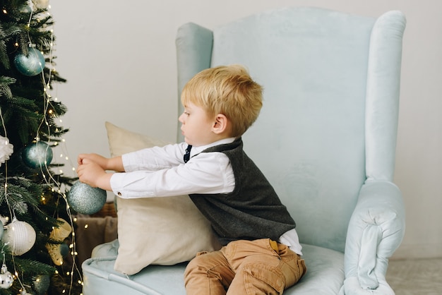 Sweet Caucasian toddler sitting in an armchair and decorating beautiful Christmas Tree