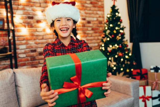 sweet christmas girl showing gift box to camera with cute smile. little child in santa hat sharing present near the decorated xmas tree with colorful packages under. beautiful laughing kid lifestyle.